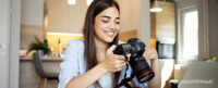 Woman smiling while holding DSLR camera and sitting t a desk in front of a laptop.