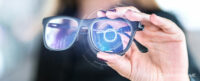 Woman holding a pair of black-rimmed glasses up to camera with blue digital graphic displayed over one of the lenses. Image used to illustrate the SmartFrame article: ‘Smart glasses: exciting or terrifying?’